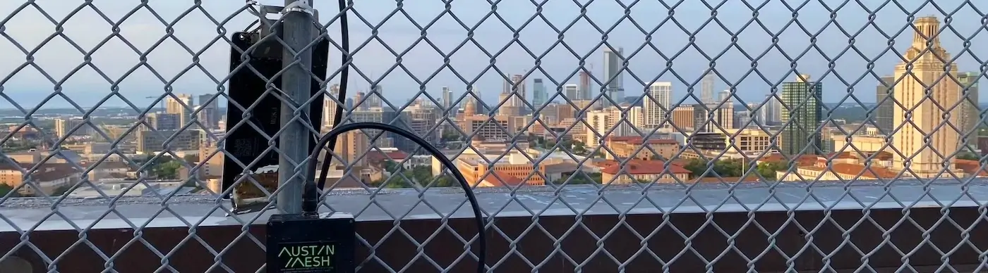 View looking out from the UT PMA building showing the Austin City Skyline to the South behind chain link fencing with a Meshtastic node that has a Austin Mesh sticker on it in the foreground